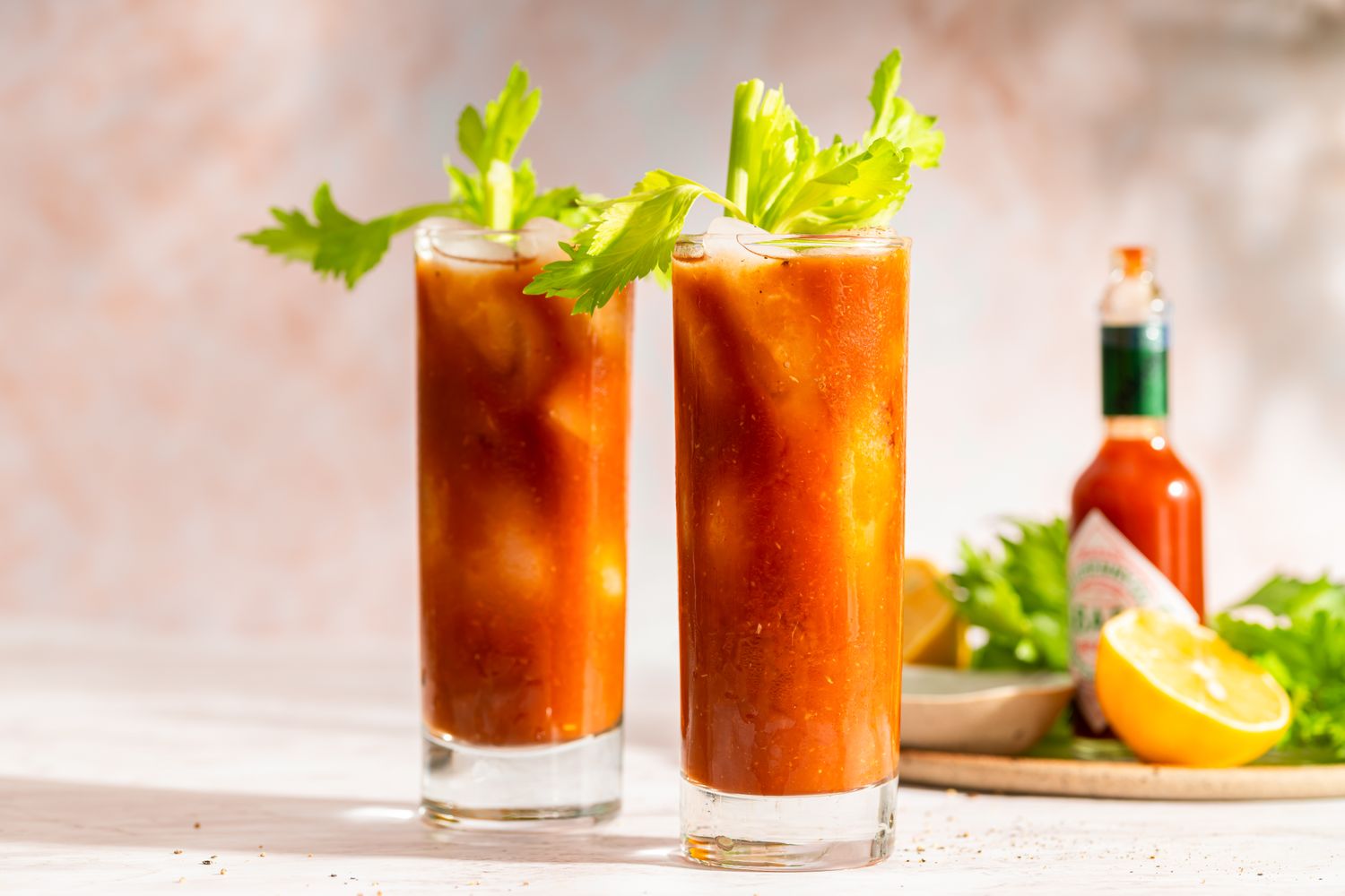 Bloody Mary cocktails garnished with celery with hot sauce and lemon slices in the background