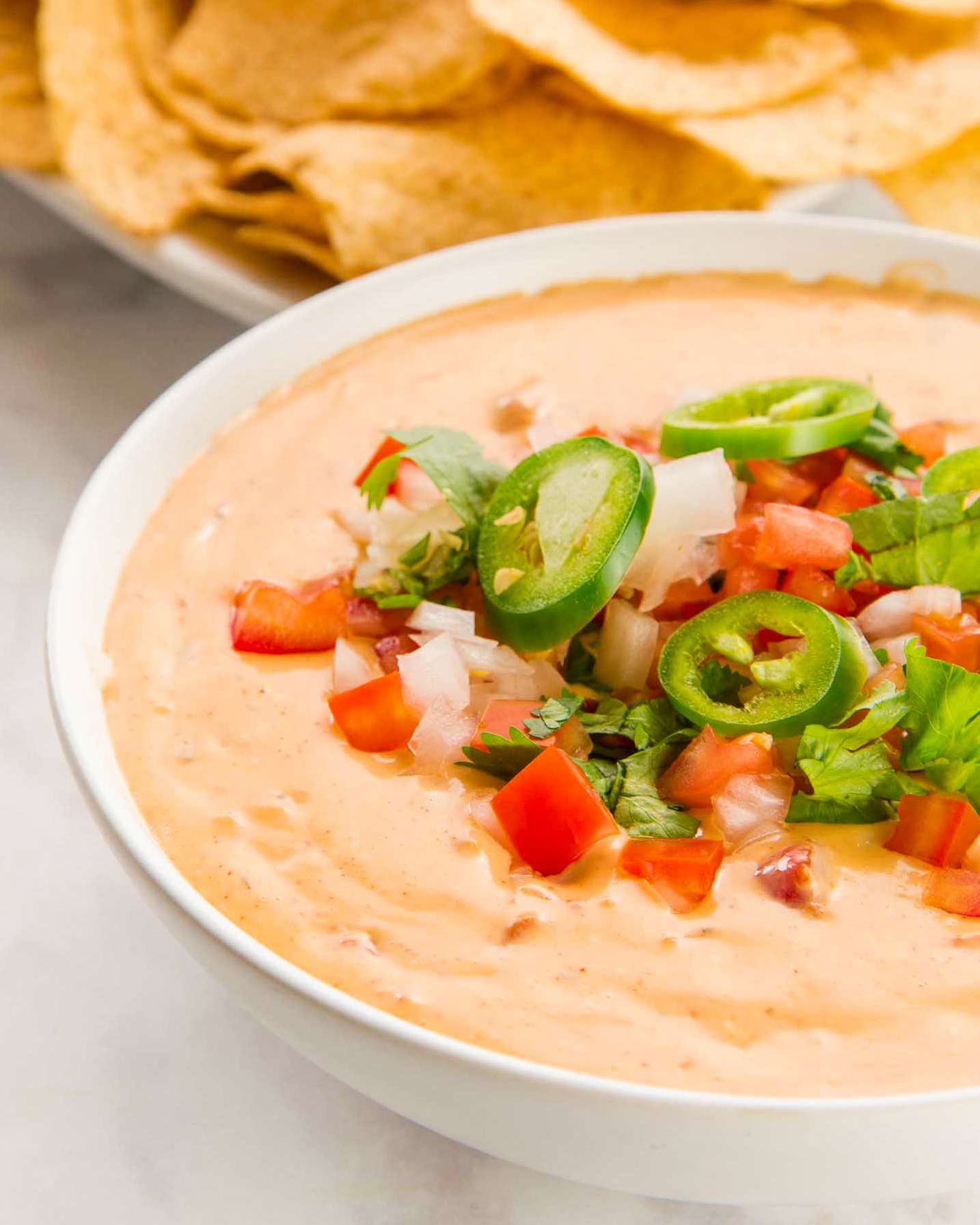 Bowl of queso dip topped with diced tomatoes onions jalapeo slices and cilantro with tortilla chips in the background
