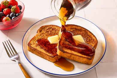 French toast with butter and syrup being poured accompanied by a bowl of berries and a fork on the side