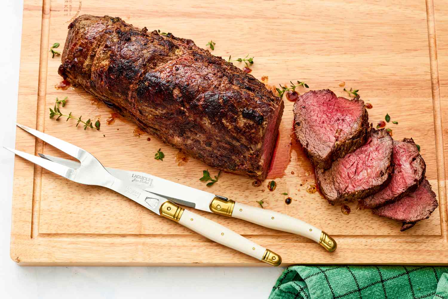 Oven Roasted Fillet of Beef on a Cutting Board With a Few Slices Laying on the Cutting Board Next to the Fillet. Next to the Meat, the Carving Utensil on the Cutting Board and a Green Kitchen Towel on the Counter