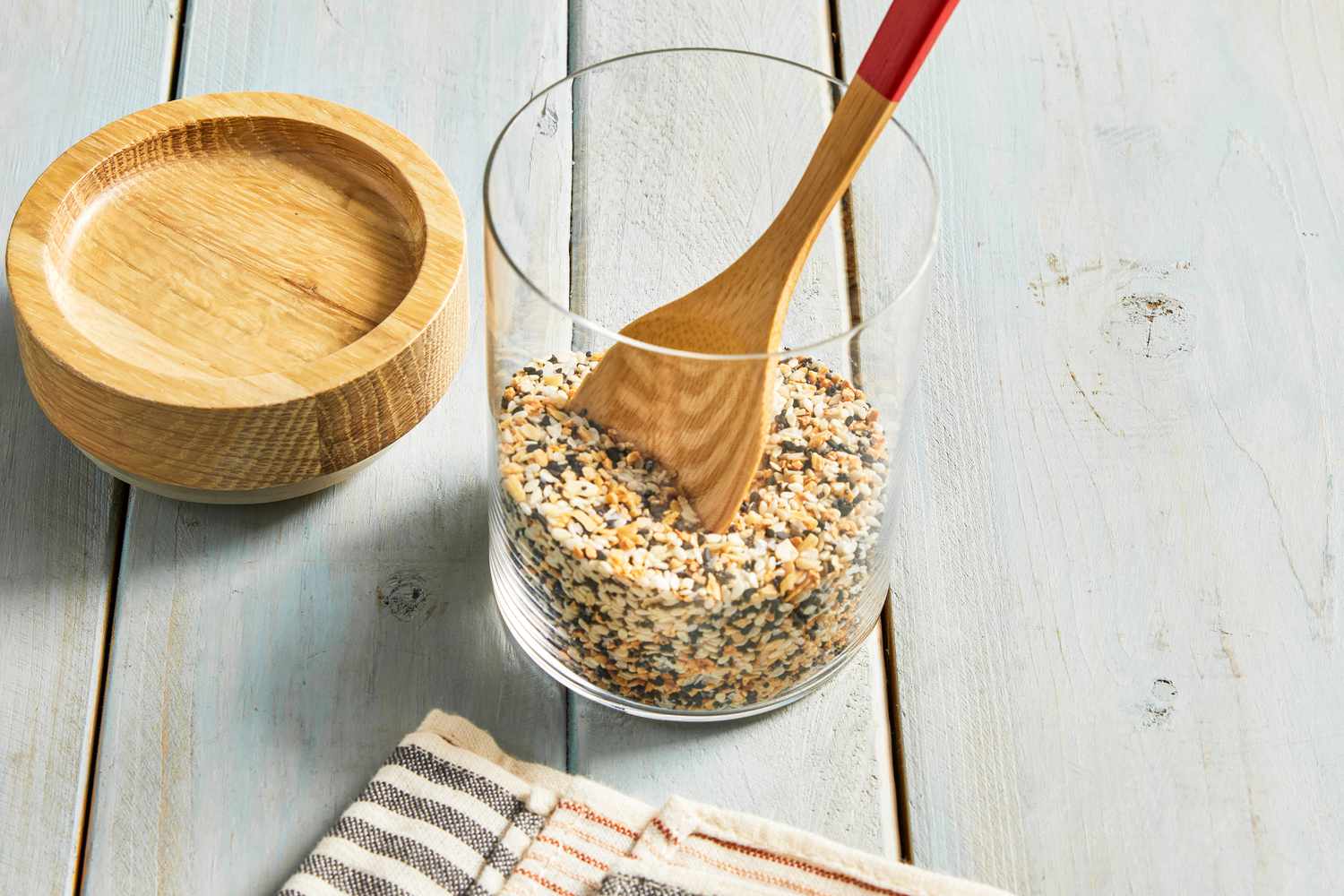 Everything Bagel Seasoning in a Tall Cylindrical Glass Jar With a Wooden Spoon In It, Next to a Wooden Circular Top and a Black and White Stripped Kitchen Towel