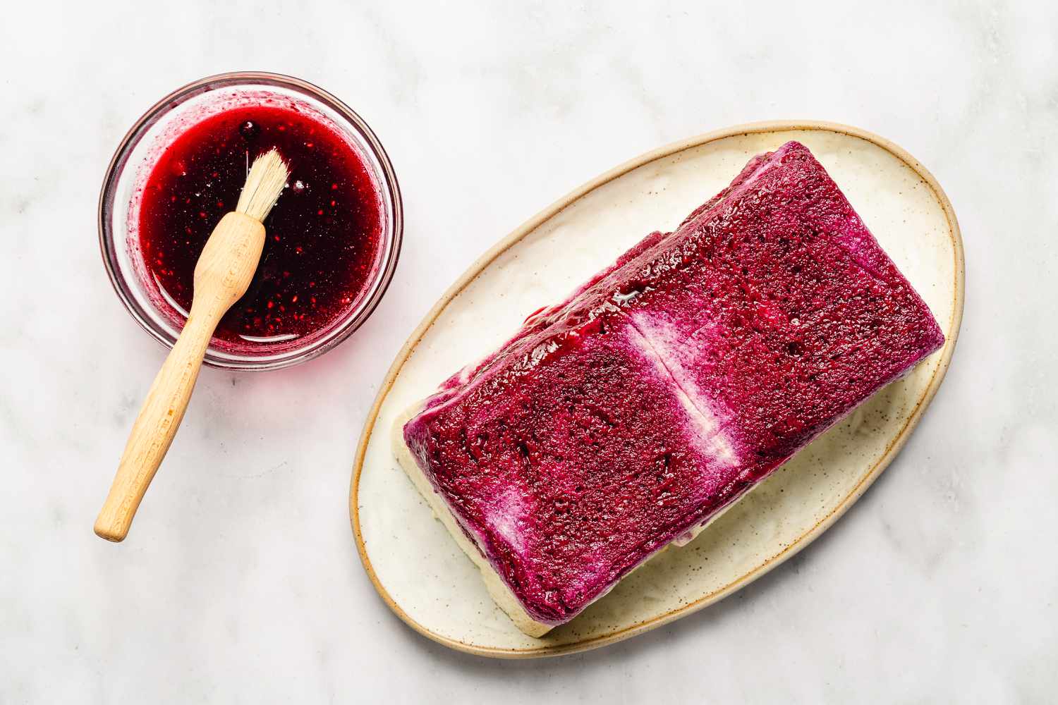 Overhead view of a bowl of berry juice and brush next to a platter with the un-molded pudding after brushing with juice for Easy British Summer Pudding recipe