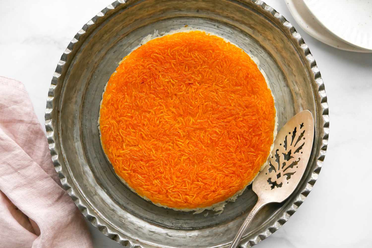 Tahdig on an Ornate Silver Platter with a Serving Spoon and Next to It, a Table Napkin and Stack of Plates