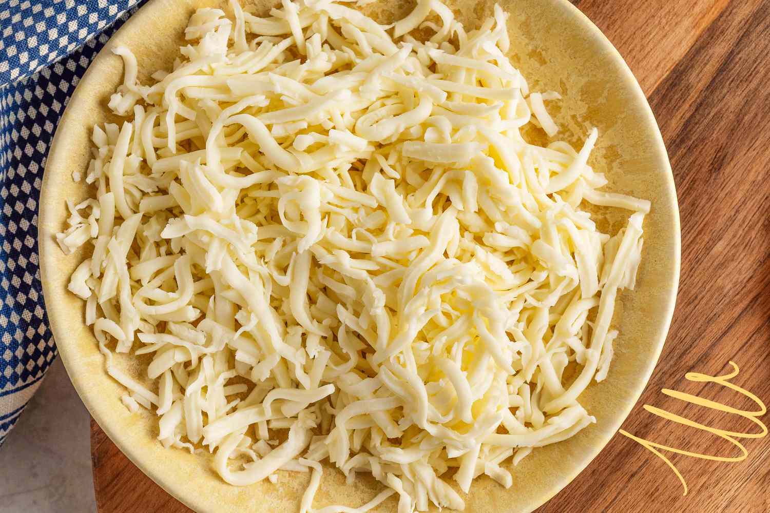 Photo of a bowl of shredded cheese and a blue and white checkered kitchen towel on a wooden tray with yellow illustrations