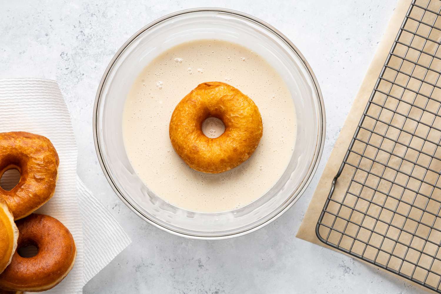 Glazing Process (L to R): Paper Towel Lined Plated with a Stack of Donuts, a Donut Dipped into a Bowl of Glaze. and a Wire Rack with Parchment Paper under It 