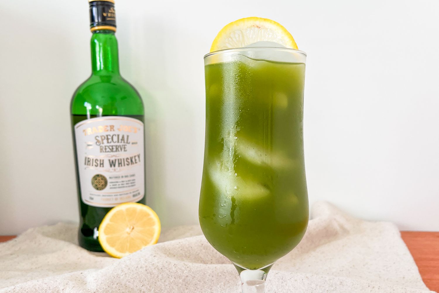 A green cocktail in a tall glass garnished with lemon and a bottle of whiskey in the background