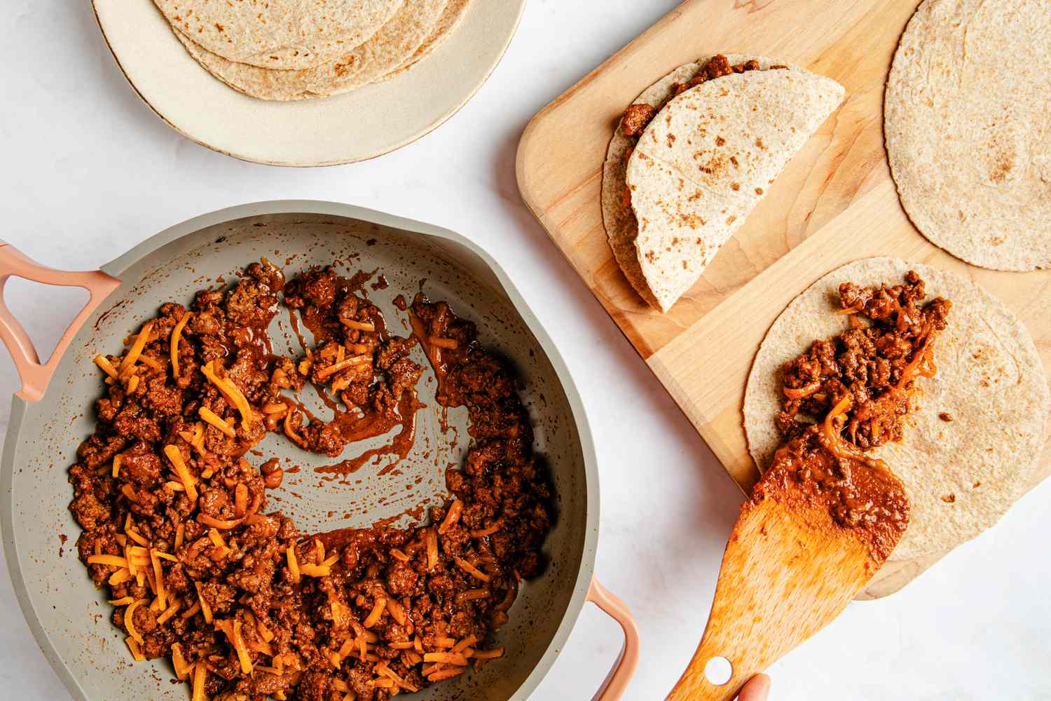 Baked taco fillings spooned onto tortillas and folded in half on a wooden board next to a plate with more tortillas and a pot with more filling