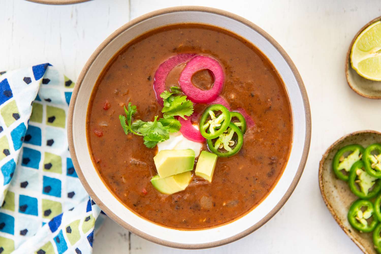Overhead shot of a bowl of black bean soup, topped with avocado cubes, jalapeño slices, pickled onion and and cilantro