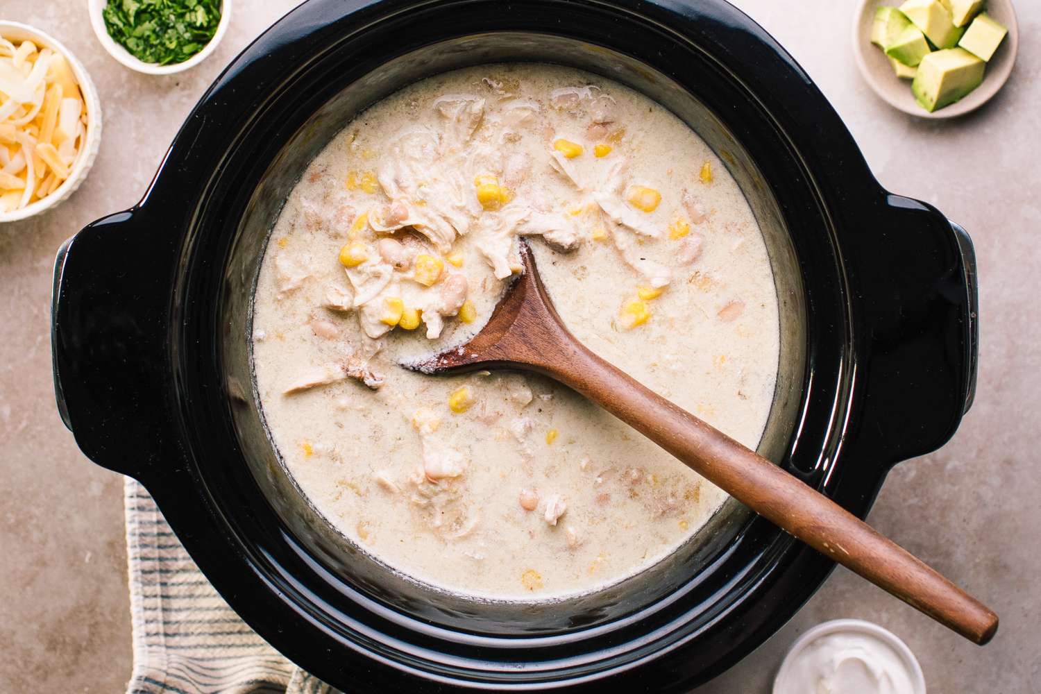 A slow cooker filled with chicken enchilada soup a wooden spoon and garnishes on the side