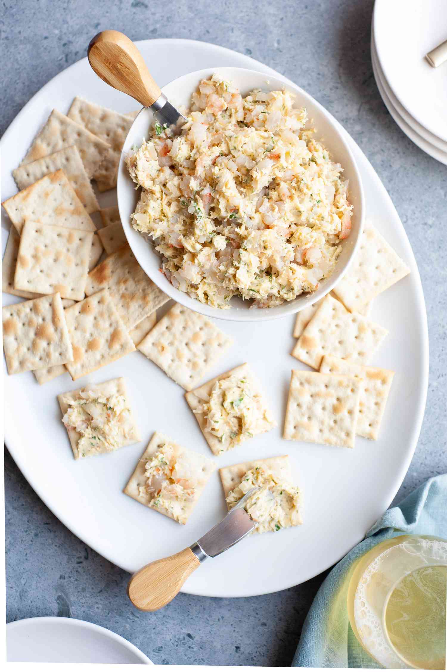 Bowl of Shrimp Butter with a Cheese Knife, Sitting on a Platter of Crackers (Some of Them Are Smeared with Shrimp Butter) at a Table Setting with a Glass of Wine, Stack of Plates, and a Kitchen Linen