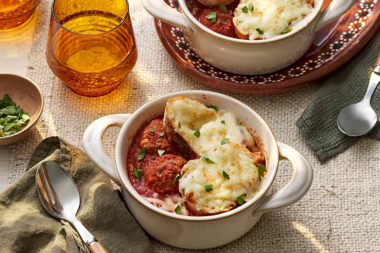 Bowl of meatball sub soup in a pot-like ramekin at a table setting with spoons, table napkins, a bowl of parsley, glasses of water, and another serving of meatball soup in a ramekin