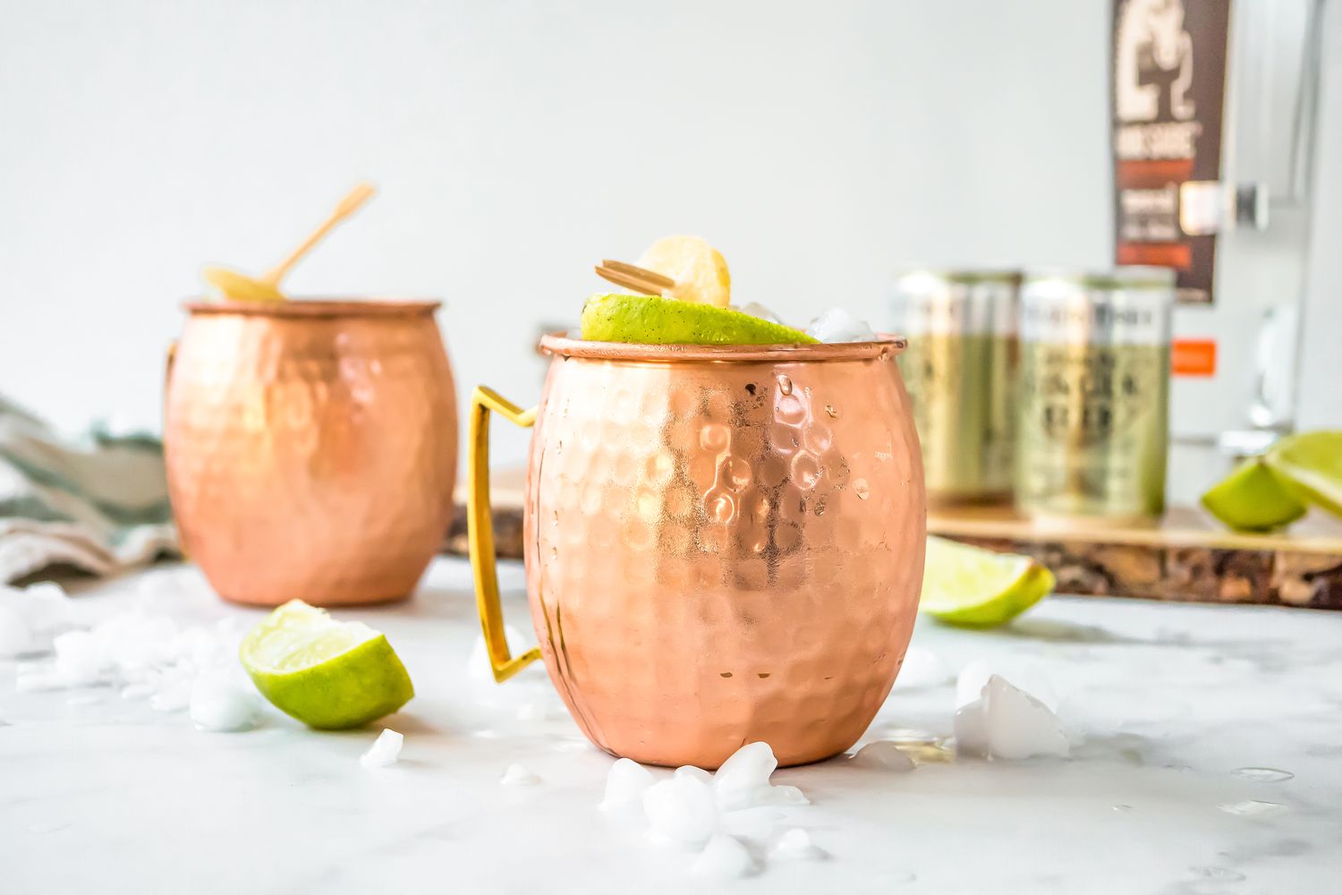 Mezcal Mule in Two Copper Mugs Surrounded by Ice and Limes. More Ingredients in the Background