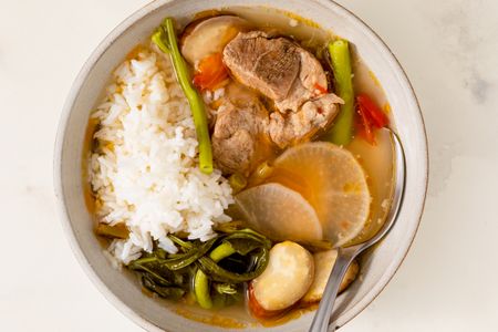 A bowl of pork sinigang served with rice and vegetables including radish green beans and eggplant with a spoon on the side