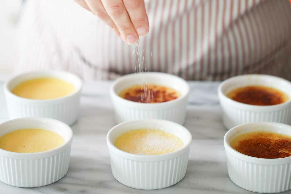 Sugar being sprinkled over the top of six crème brûlées in ramekins
