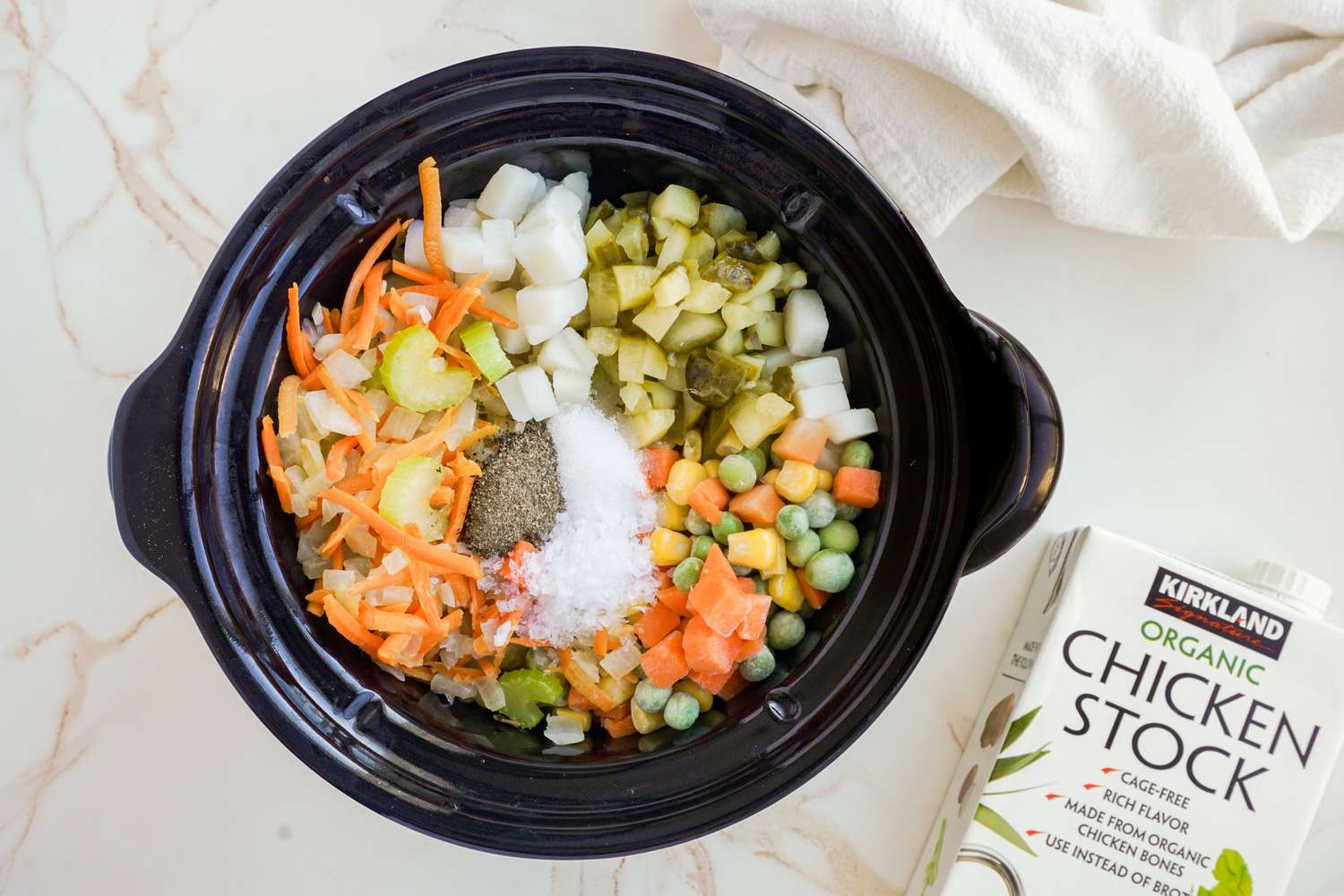 Ingredients in a black slow cooker including various vegetables and spices with a carton of chicken stock nearby