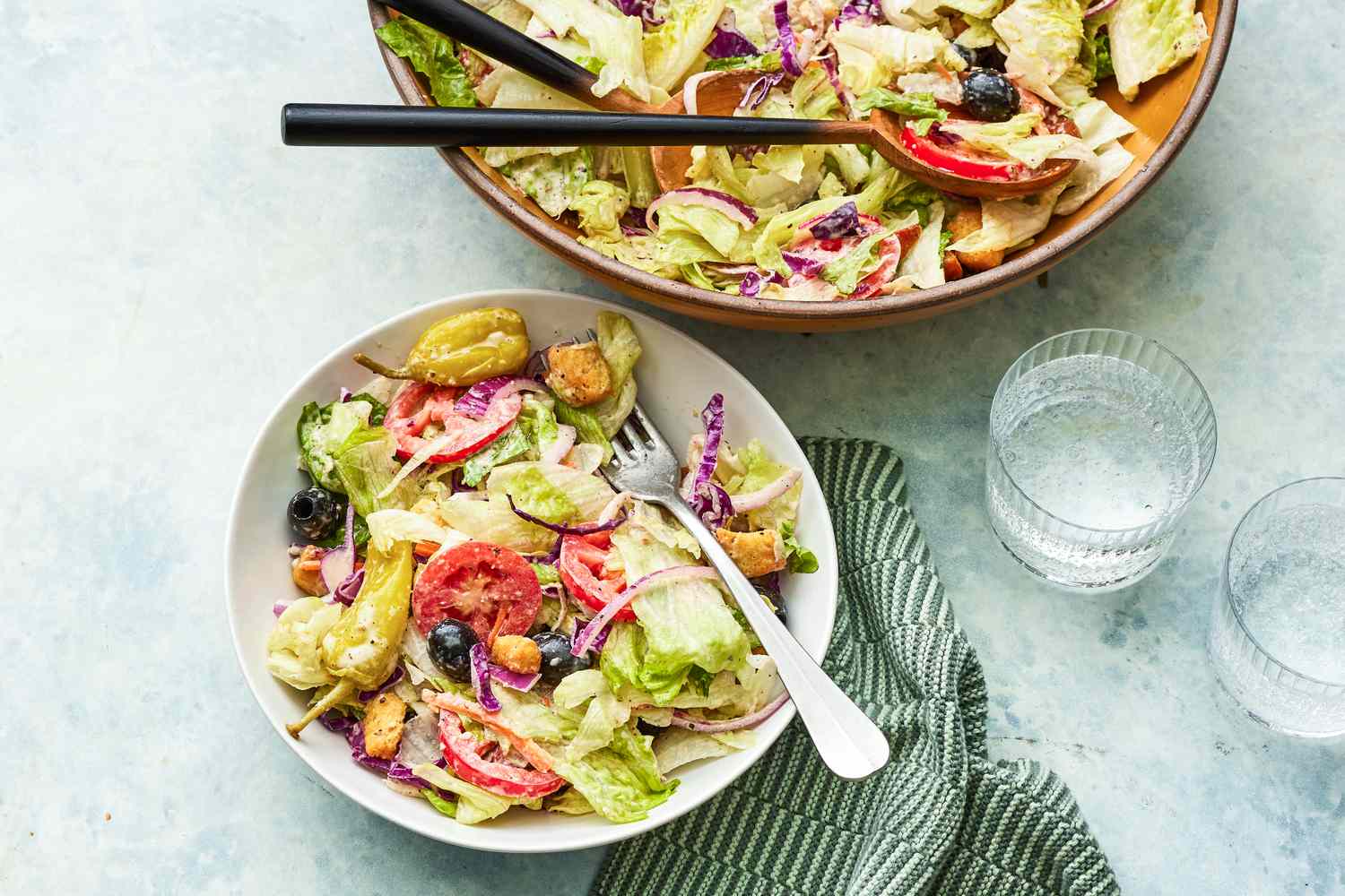 A Serving of Olive Garden Salad Next to a Larger Bowl of Olive Garden Salad With Serving Utensils, and on the Counter Next to it, a Glass With Salad Dressing and a Green Striped Table Napkin