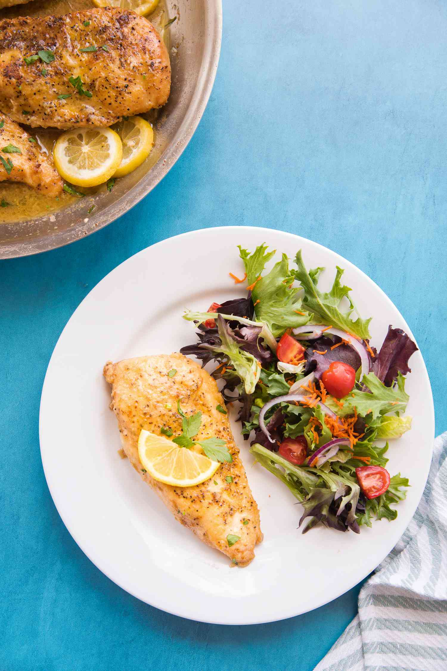 On a Plate, a Serving of Lemon Pepper Chicken Topped with Parsley and a Slice of Lemon and Served with a Side Salad, and Next to the Plate, a Pan with More Lemon Pepper Chicken 