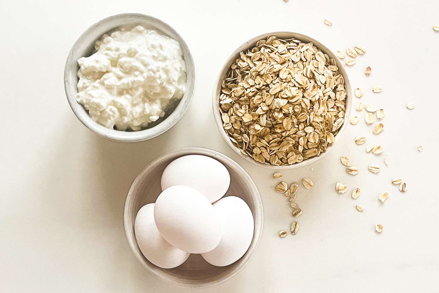 Bowls containing cottage cheese, oats, and three raw eggs arranged on a light surface