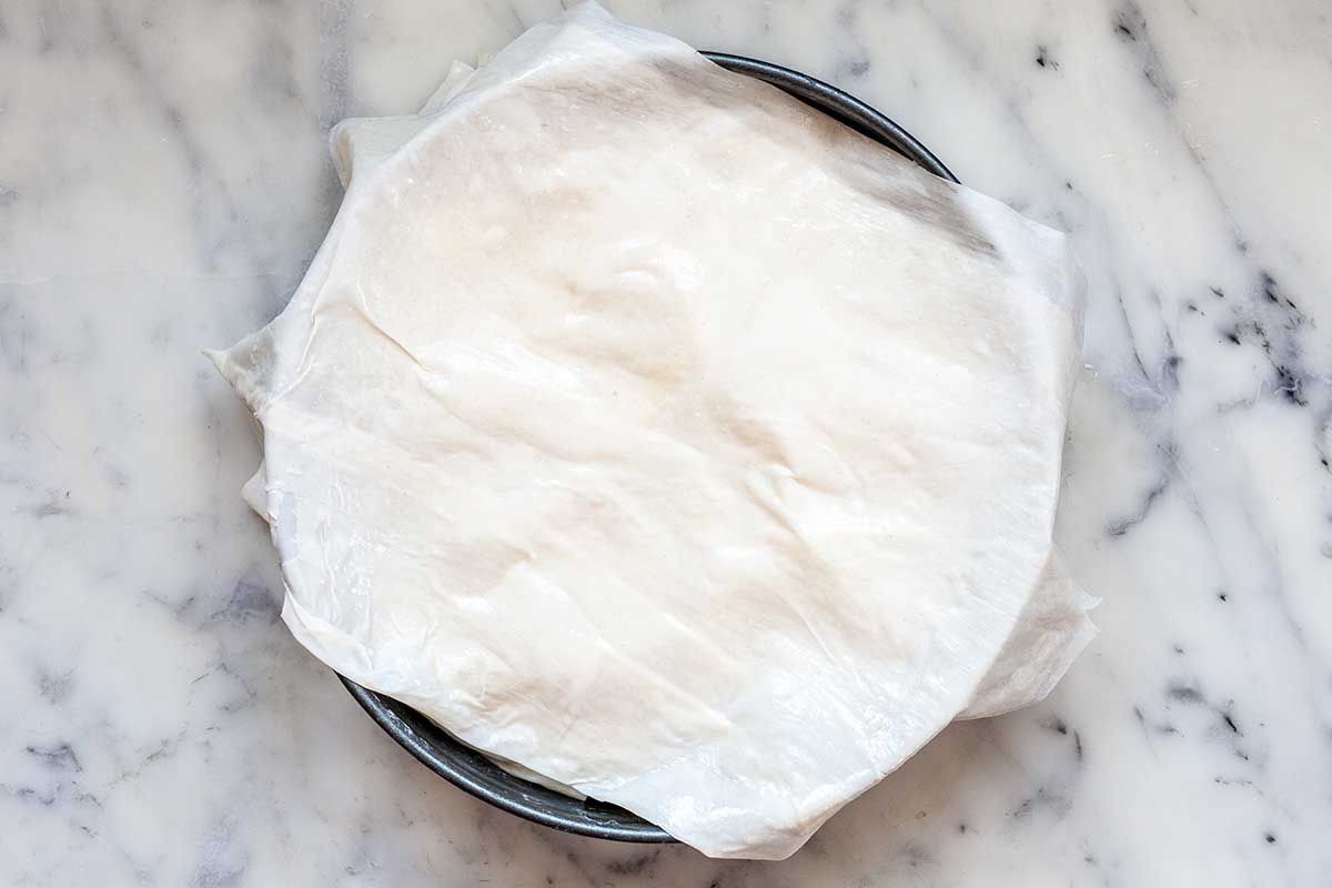 A cake pan is on a marble countertop. Unbaked phyllo dough is covering the top and overlapping the pan.
