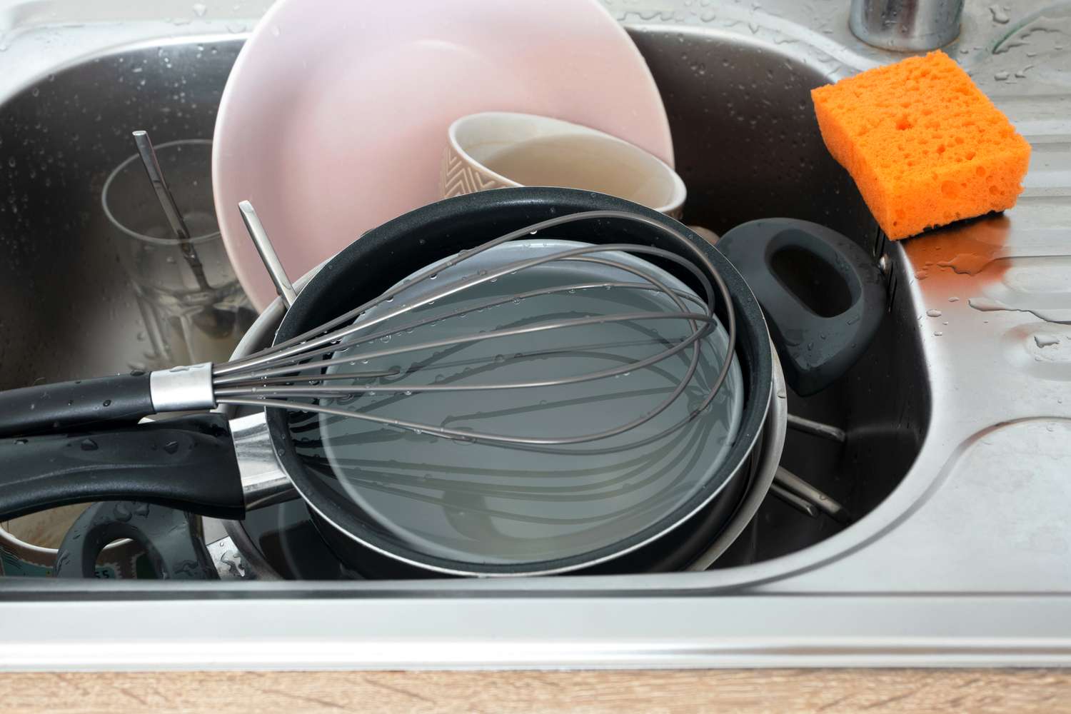 Sink containing dishes pans and cooking utensils with an orange sponge beside it