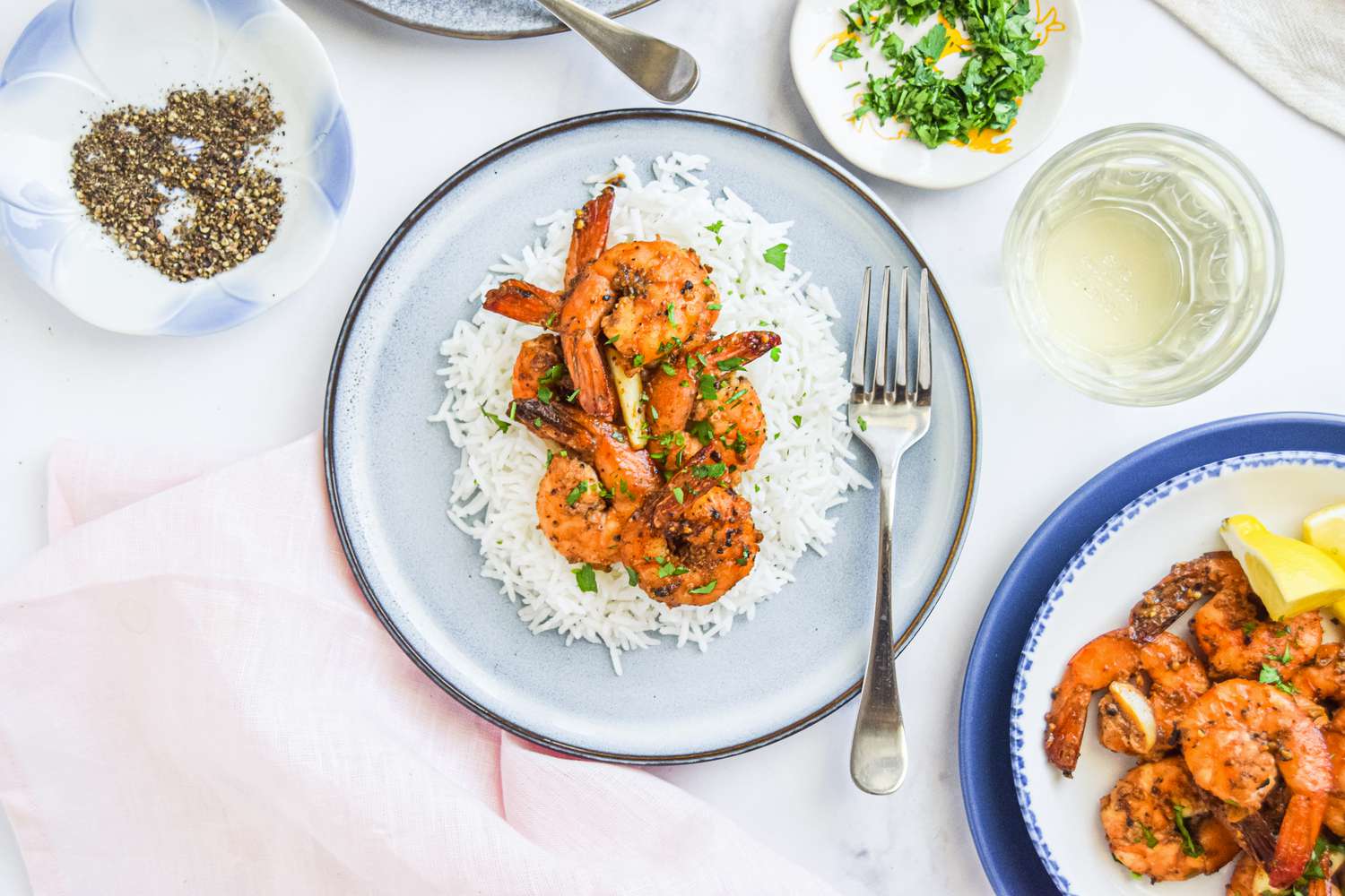 Peppery Garlic Shrimp Over Rice on a Plate Next to a Plate with More Shrimp and Lemon Wedges, a Glass, and Small Plates with Seasoning and Herbs