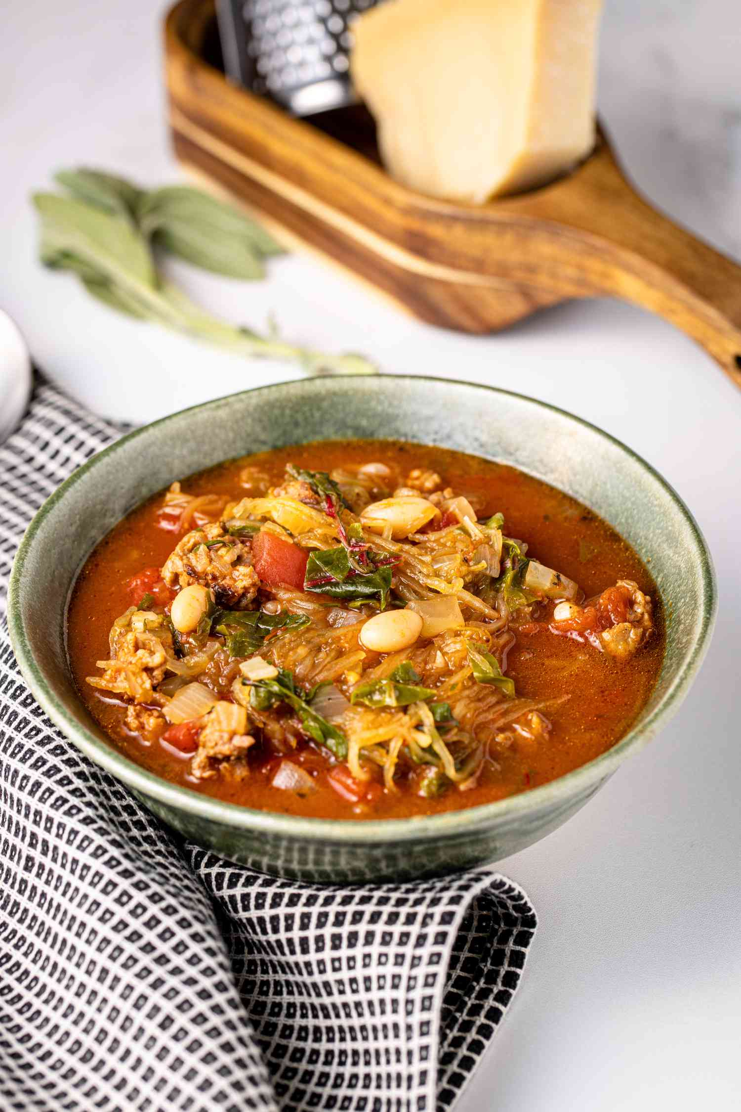 Spaghetti Squash Soup with Italian Sausage in a Bowl Next to a Kitchen Towel and a Spoon and in the Background, a Parmesan Cheese and a Grater in a Wooden Holder