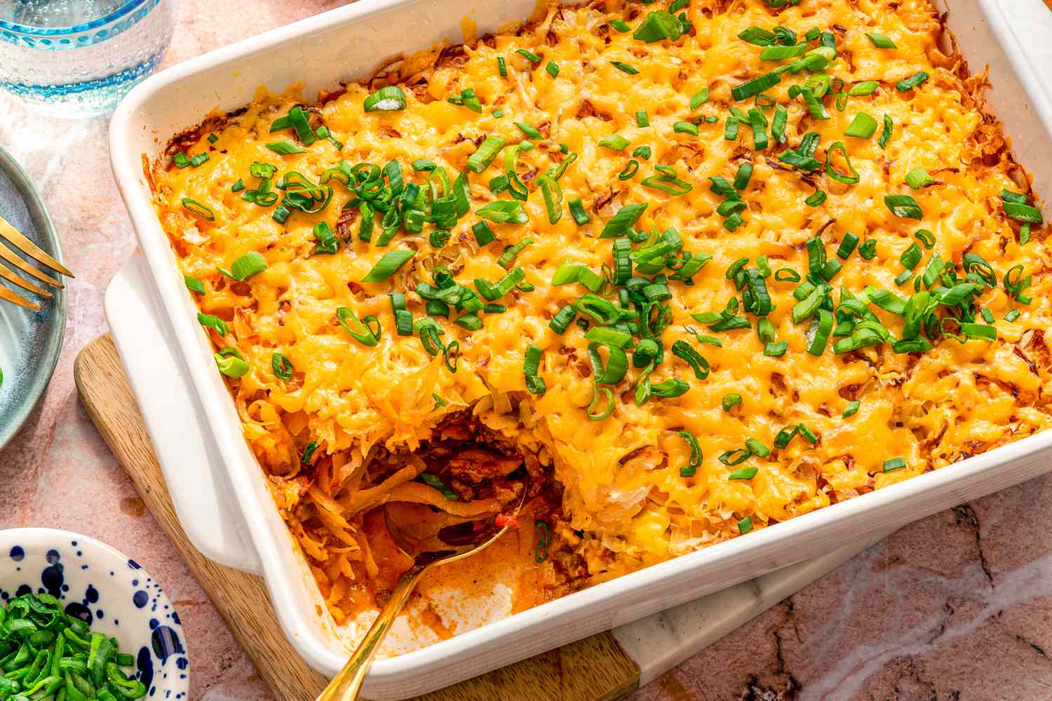 A baked casserole topped with melted cheese and chopped green onions served in a white dish with a spoon scooping from one side