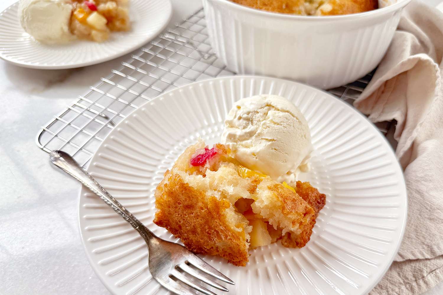 A slice of fruit cocktail cake served on a plate with a scoop of ice cream on top and a fork placed nearby