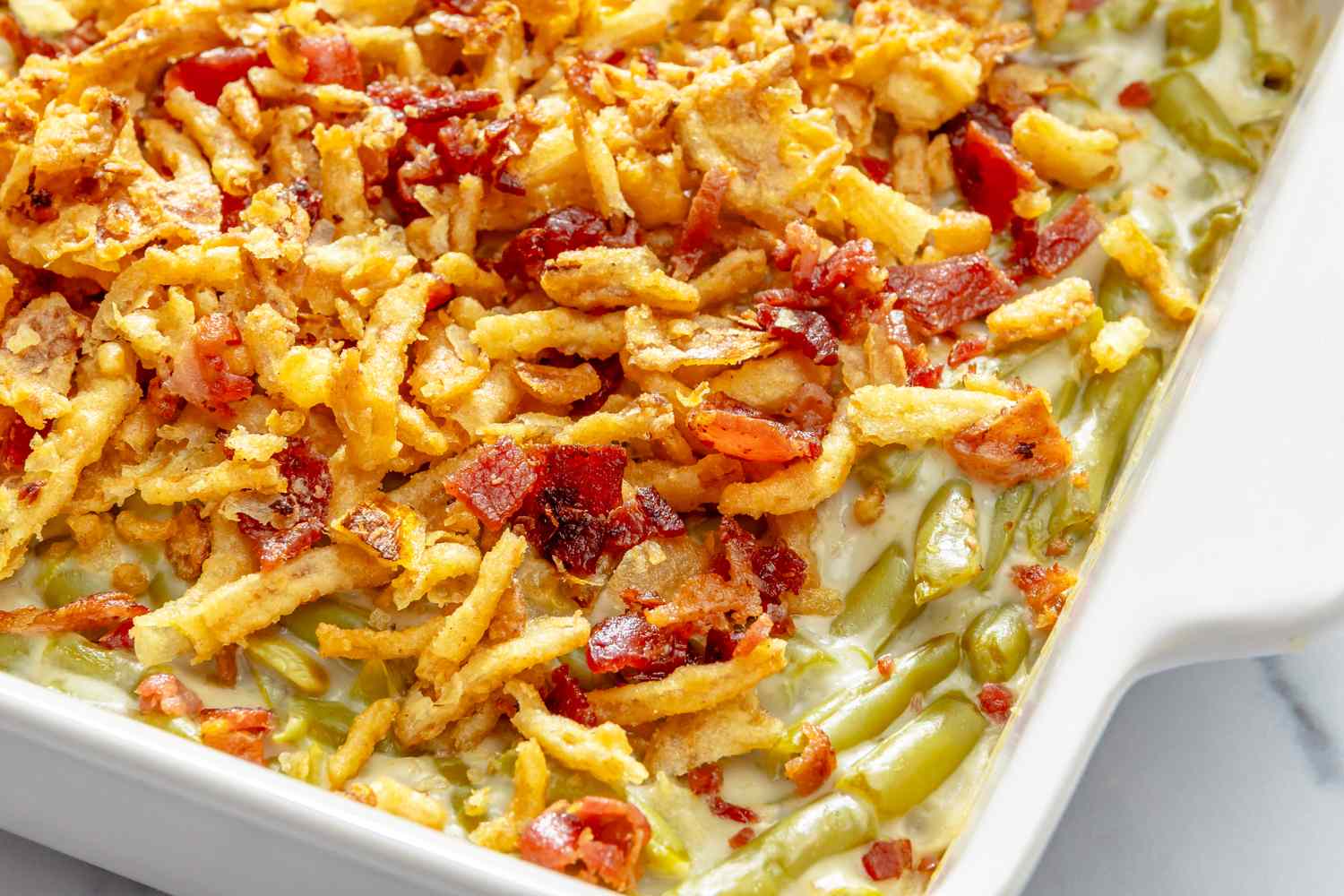 A dish of green bean casserole topped with fried onions and bits of bacon served in a rectangular white baking dish