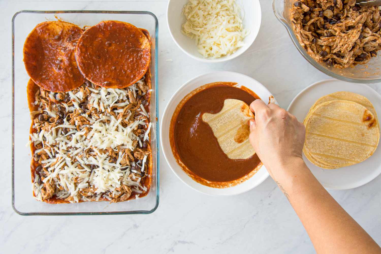 Assembling the Casserole (L to R): Casserole Dish With Layers of Casserole Ingredients, a Tortilla Doused in a Bowl of Red Enchilada Sauce, Bowl of Shredded Cheese, Bowl of Seasoned Chicken and Beans, and a Plate With a Stack of Tortillas for Chicken Enchilada Casserole Recipe