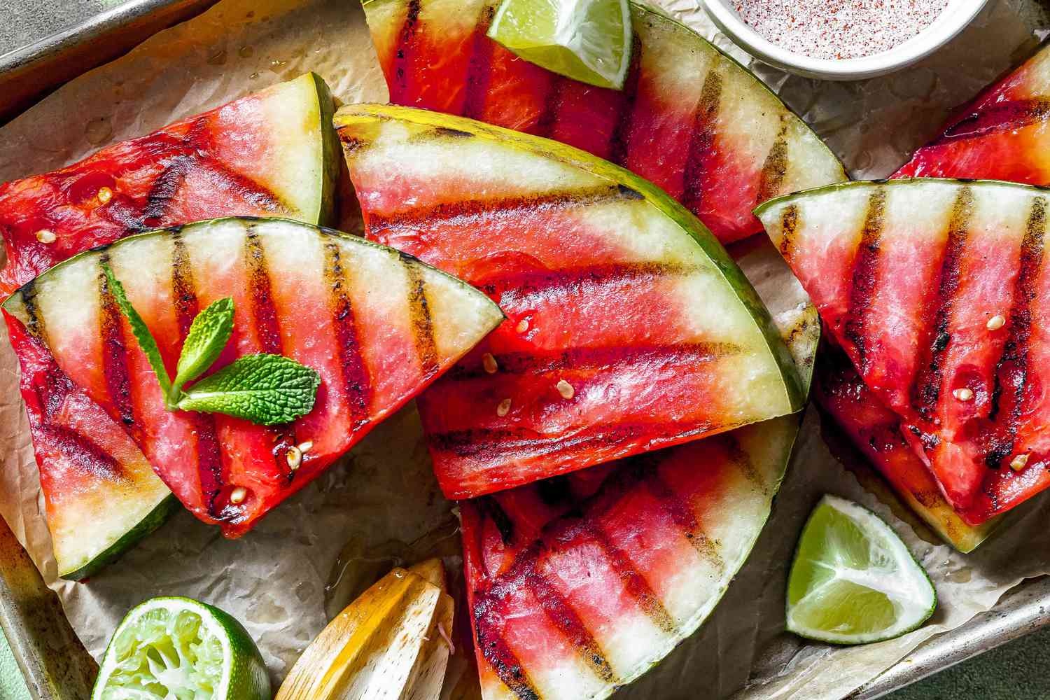 Triangles of grilled watermelon with small bowl of spice mix, sprig of mint and lime wedges alongside