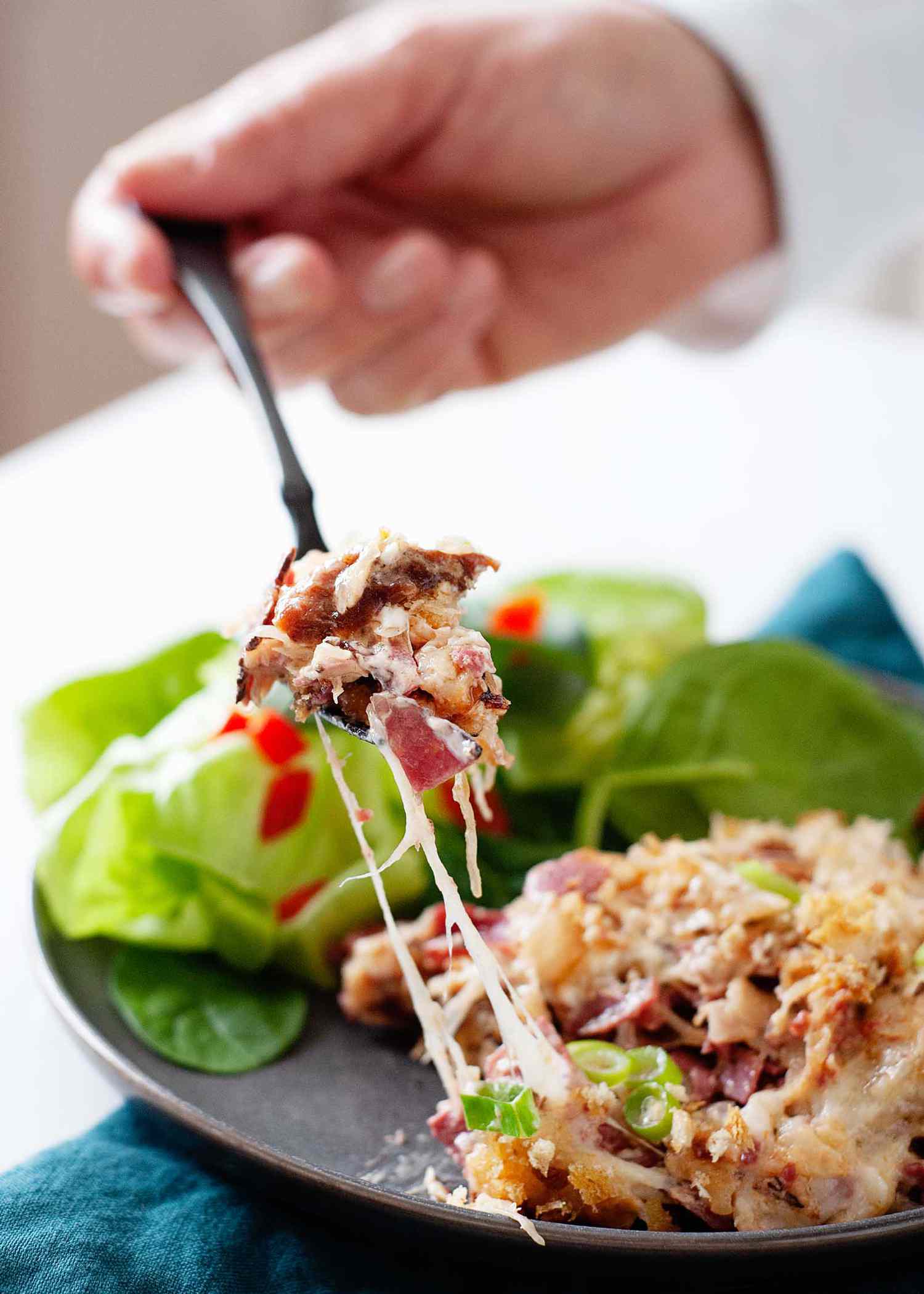A scoop of reuben casserole is on a matte grey plate with a teal linen underneath. A forkful of cheesy casserole is lifted above the scoop on the plate.