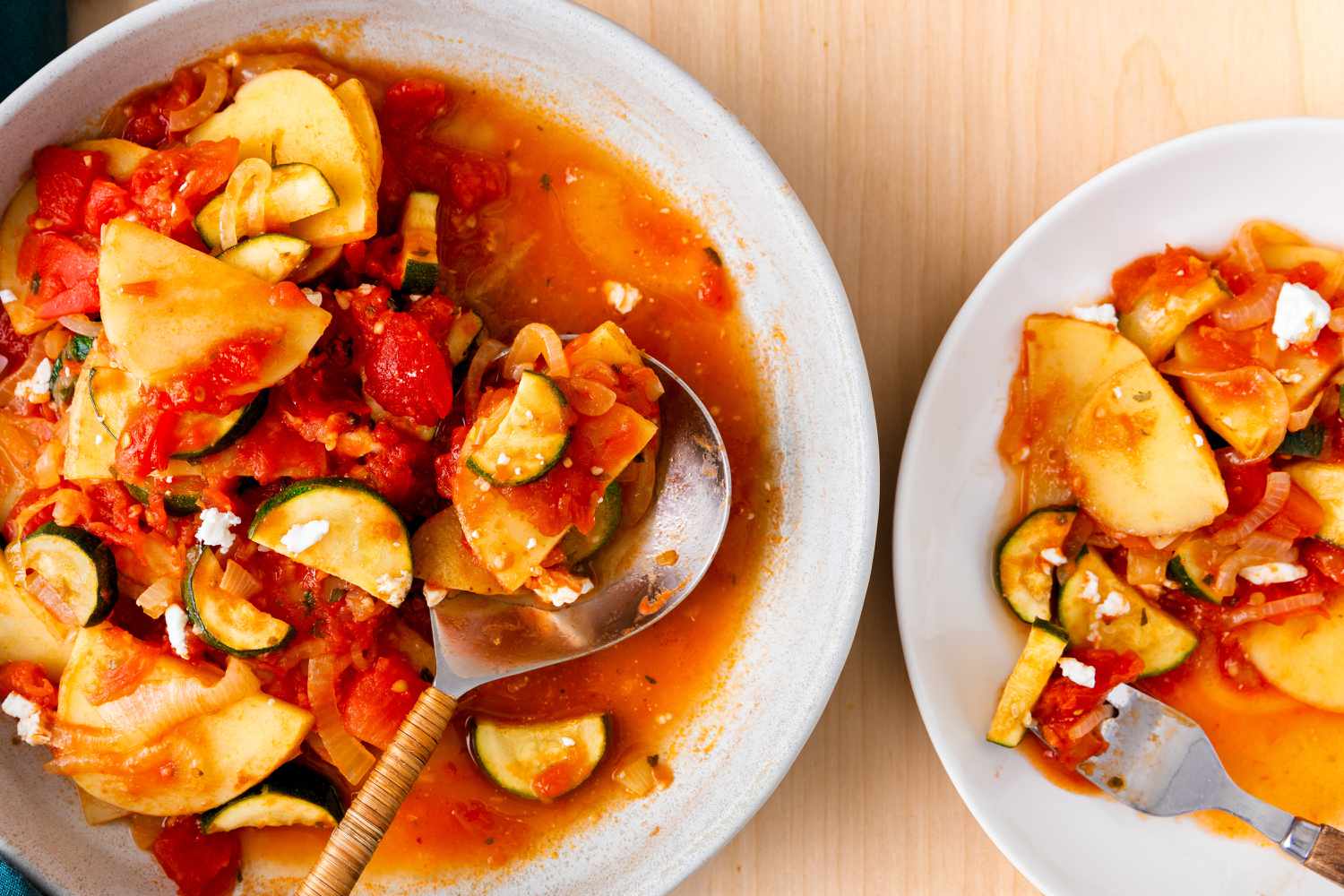 Overhead view of a white bowl of Zucchini Con Patate with a spoon in it on a table next to a smaller bowl of with a fork