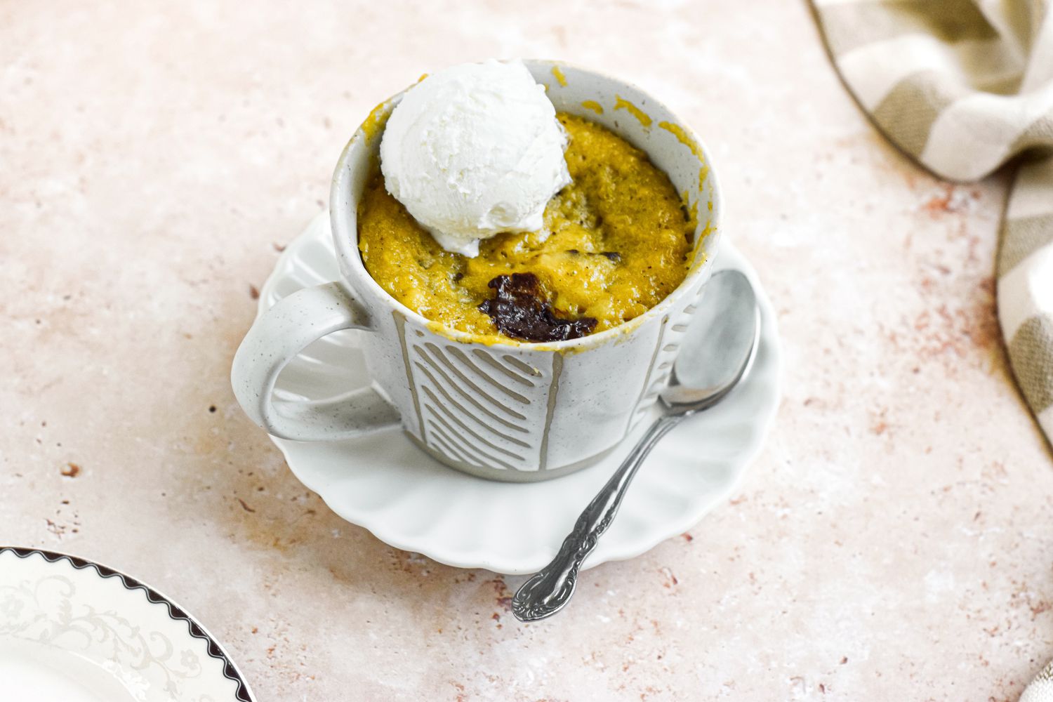 Mug of Banana Bread Chocolate Chunk Mug Cake Topped with a Dollop of Ice Cream on a Saucer With a Spoon Next to a Kitchen Towel