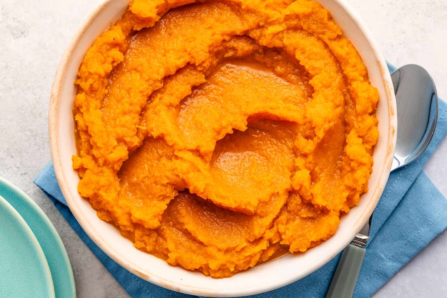 Overhead view of a white bowl of butternut squash resting on a blue table napkin next to a spoon
