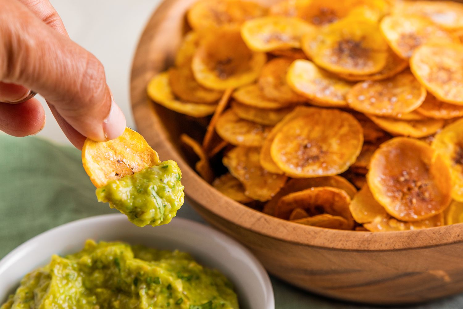 Hand Dipping a Plantain Chip in a Bowl of Guacamole, and in the Background, a Large Bowl Filled With Plantain Chips