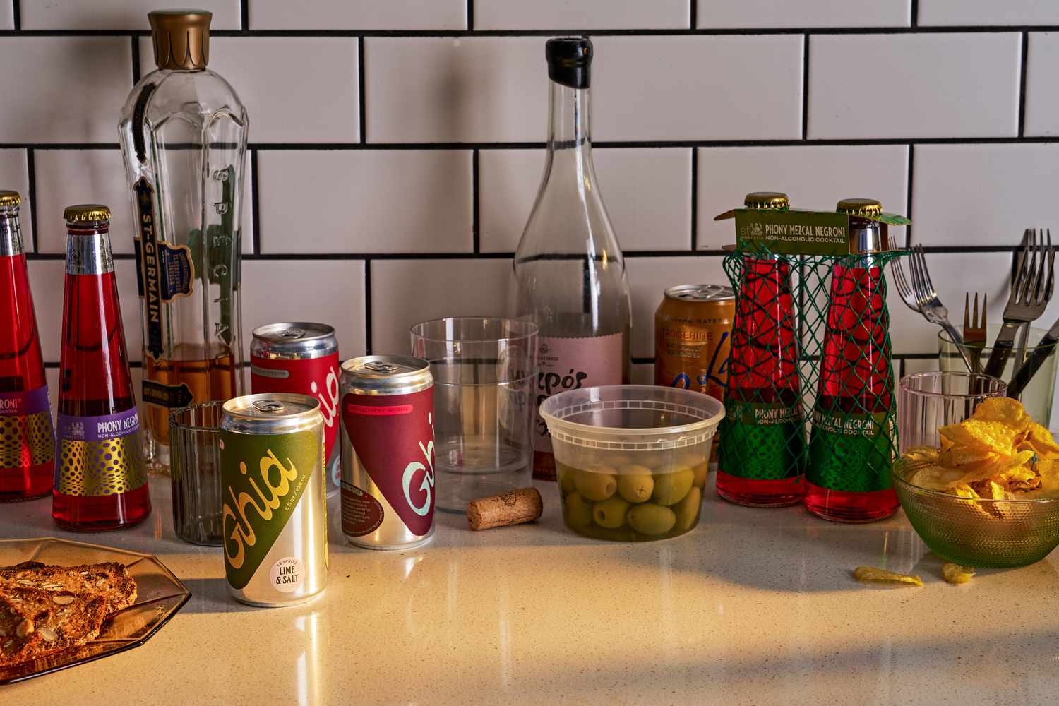 A kitchen countertop with a mix of snacks, alcoholic beverages, and non-alcoholic beverages like Ghia and Phony Negroni