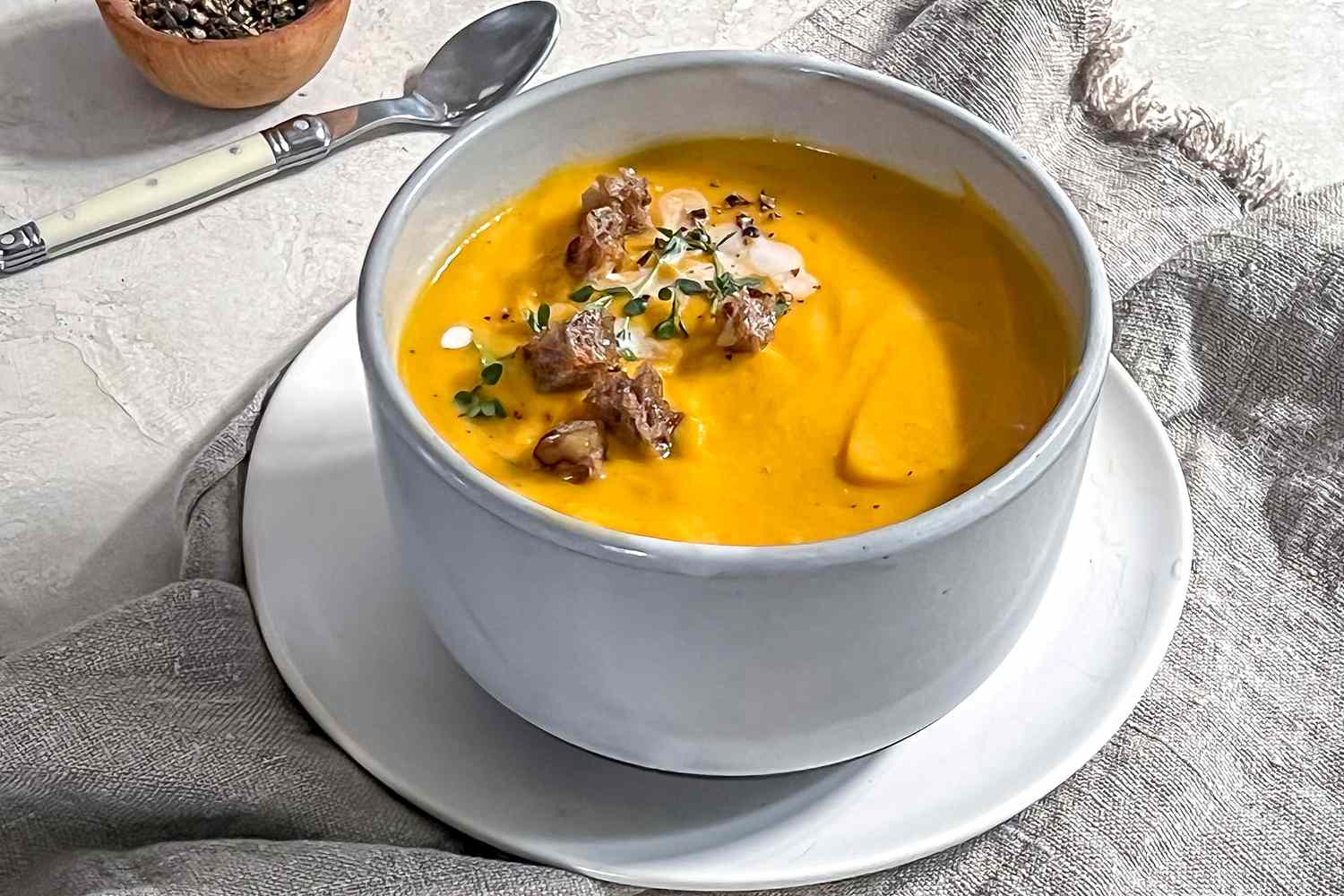 Bowl of Vegan Butternut Squash Soup Topped with Thyme, Coconut Milk, Croutons, and Cracked Pepper, Sitting on a Kitchen Linen and Surrounded by a Bowl of Cracked Pepper and a Spoon 
