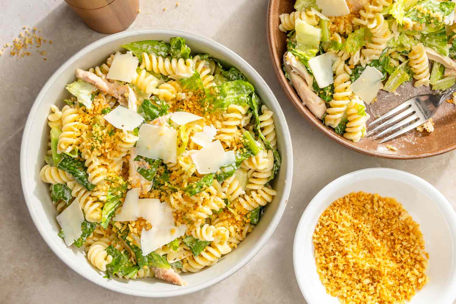 Bowl of rotini pasta salad with chicken and lettuce, garnished with shaved cheese and a dish of bread crumbs beside it