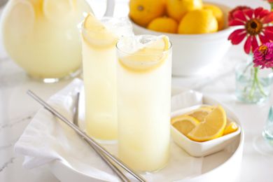 Two glasses of lemonade with lemon slices on a tray, a bowl of lemons, and a vase of flowers in the background