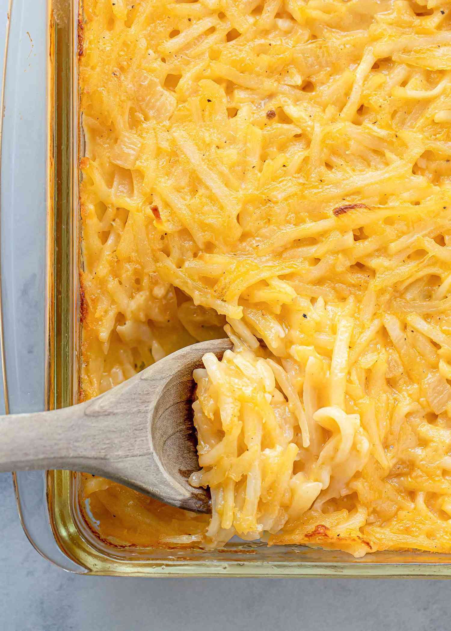 Overhead vertical view of a glass dish filled with mormon funeral potatoes. A wooden spoon is scooping some of the cheesy funeral potatoes. Only a fourth of the dish is in view.