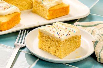 A slice of vanilla cake with frosting and sprinkles on a white plate, next to a fork and a napkin, with a tray of cake pieces in the background