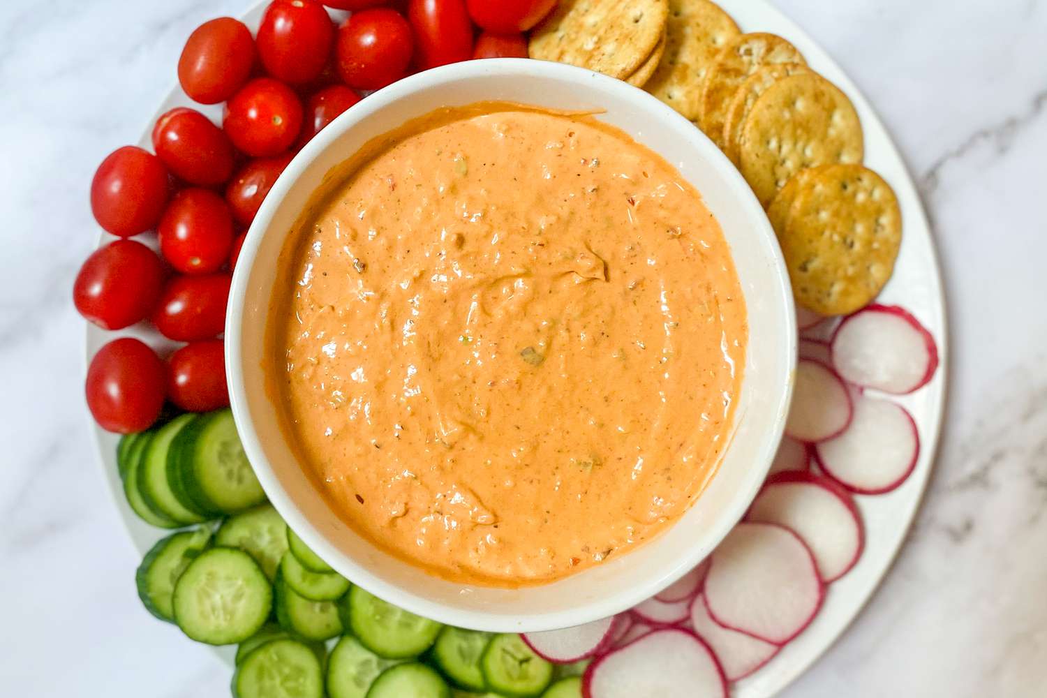 A bowl of orange dip surrounded by vegetables and crackers on a plate
