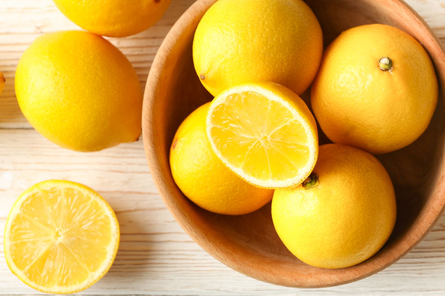 A wooden bowl with several whole lemons and one halved lemon on a light wooden surface