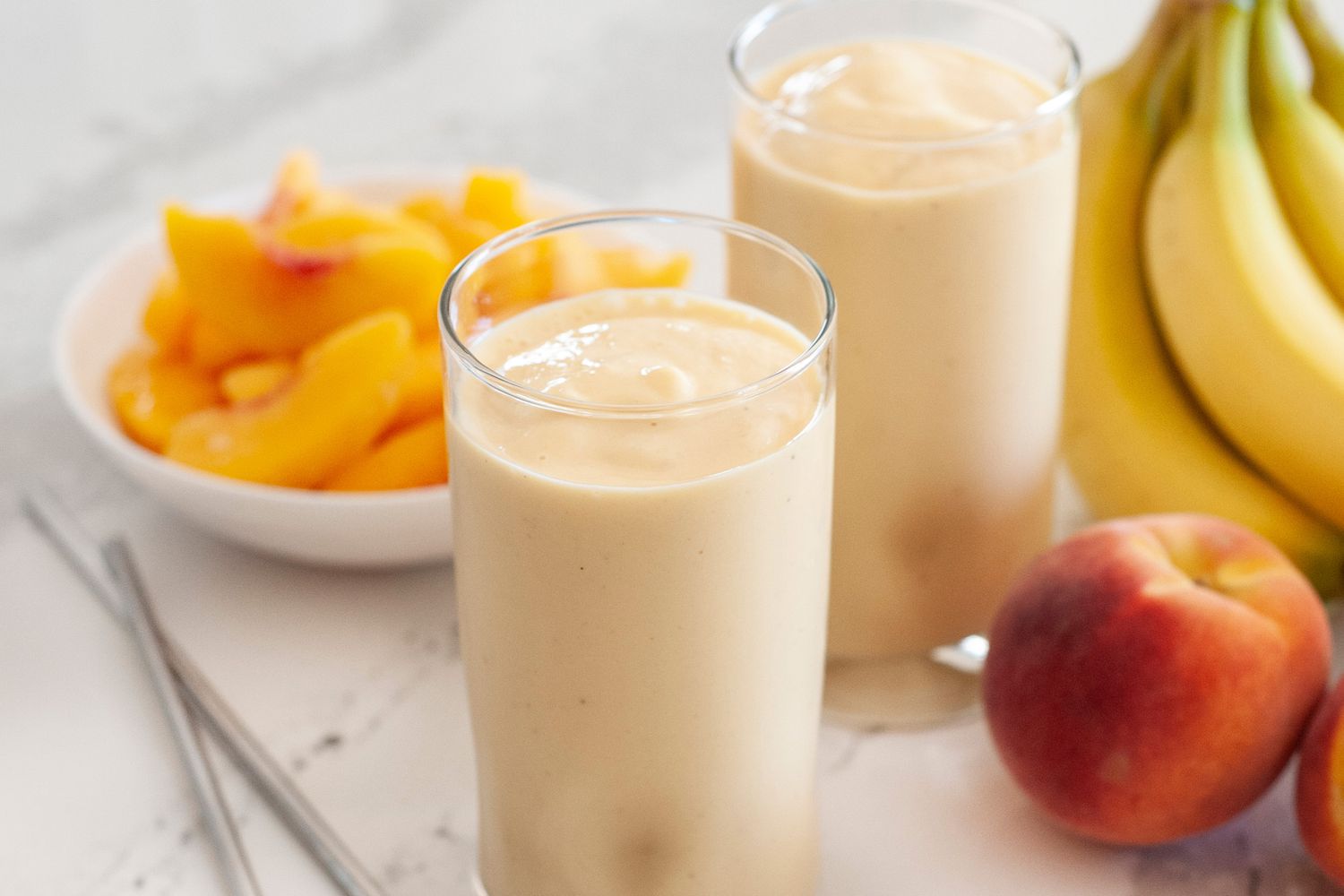 Two peach almond smoothies with whole bananas, peaches, and a plate of sliced peaches set around the smoothies.