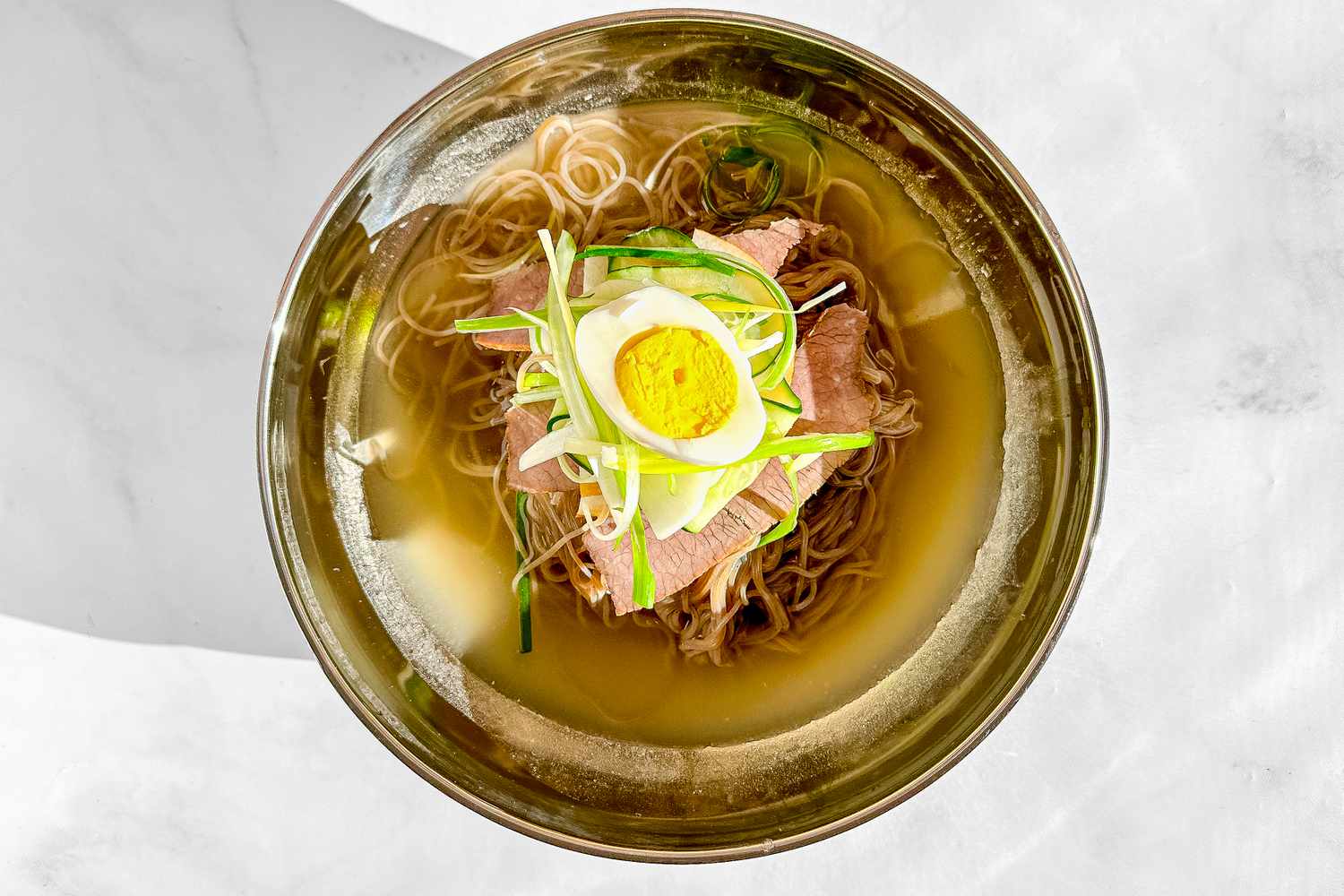 Bowl of Naengmyeon Topped With a Halved Hard Boiled Egg, Cucumber Ribbons, and Julienned Green Onions