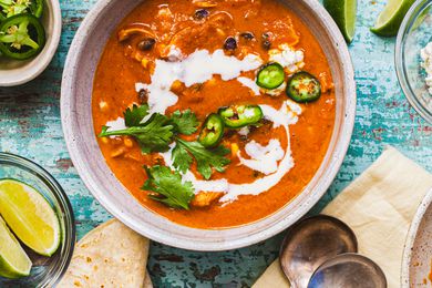 Creamy Chicken Enchilada Soup Topped with Jalapenos, Cilantro, and Crema in a Bowl at a Table Setting with Two Spoons on a Table Napkin, Bowls of Toppings (Jalapenos and Lime Wedges), and Tortillas