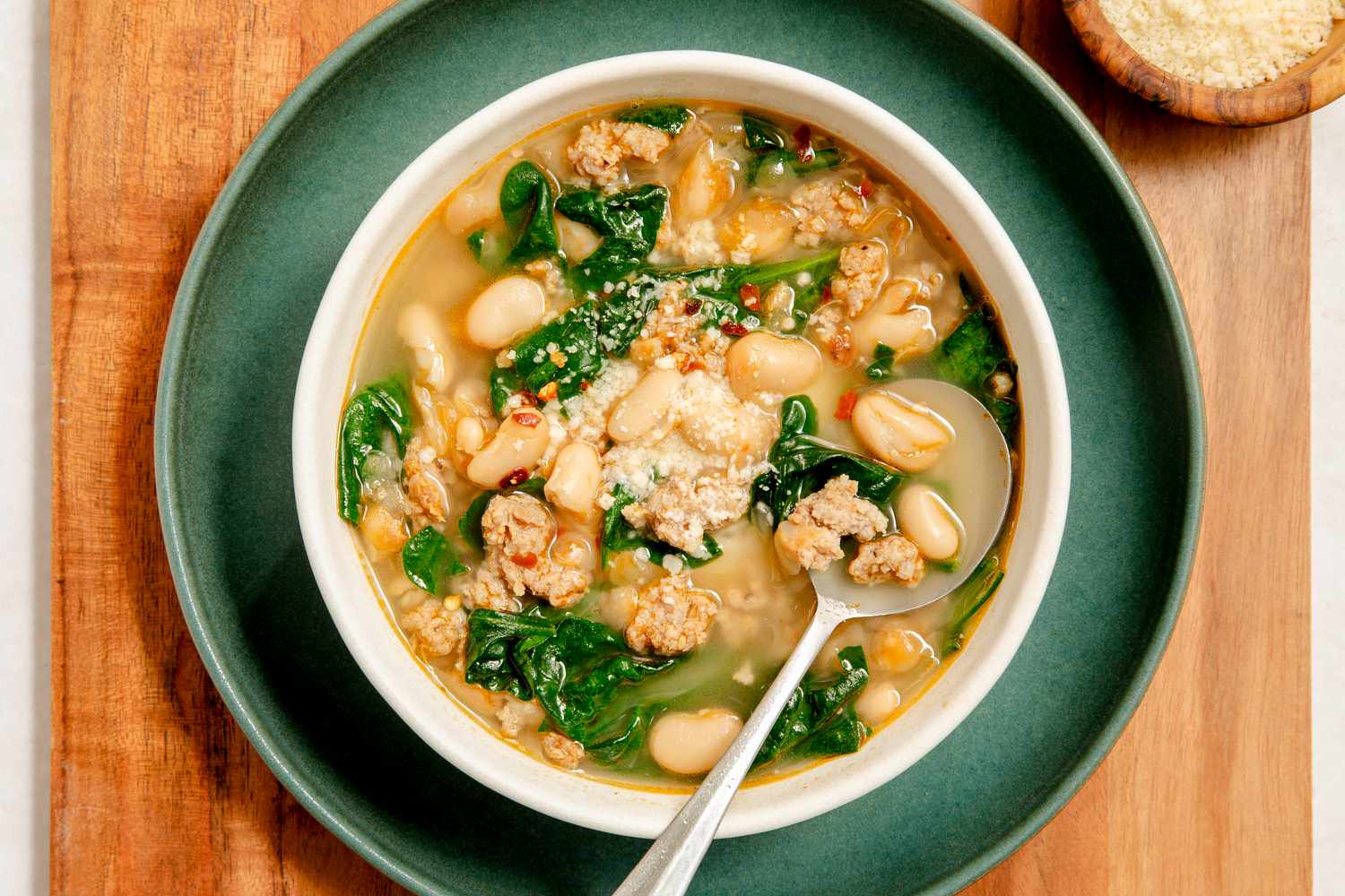 A white bowl with kale, white bean and sausage soup, places on a dark green plate on a wooden serving board