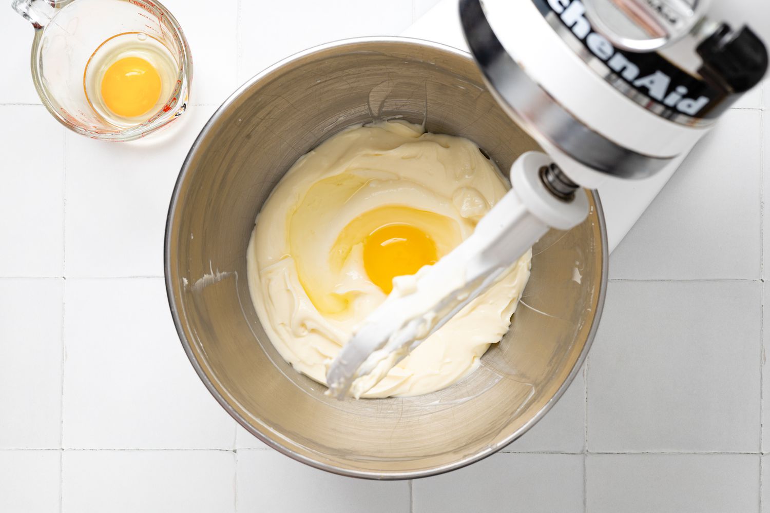 An Egg Added the Cream Cheese in a Mixer Bowl on a Stand Mixer, and on the Counter, a Pyrex Measuring Cup With Another Egg for Lemon Cheesecake Bars Recipe