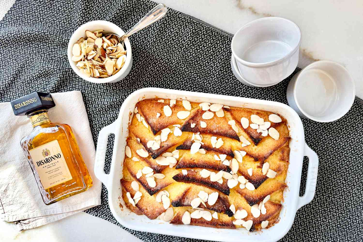 Overhead view of a baking dish of Princess Diana's favorite bread pudding along with a bottle of Disaronno amaretto liqueur and a bowl of almond slivers on a marble countertop
